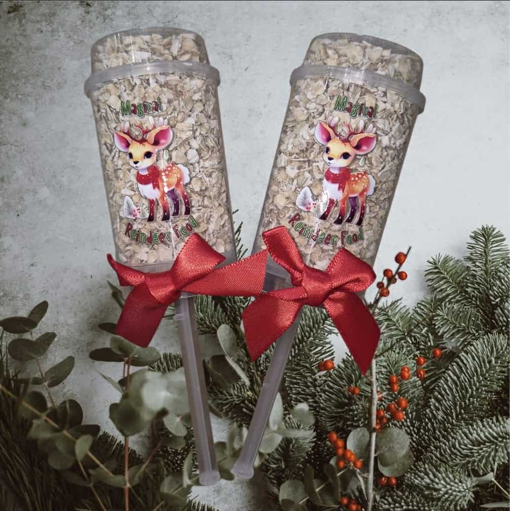 Two tumblers with deer designs and red bows, surrounded by greenery on a light background